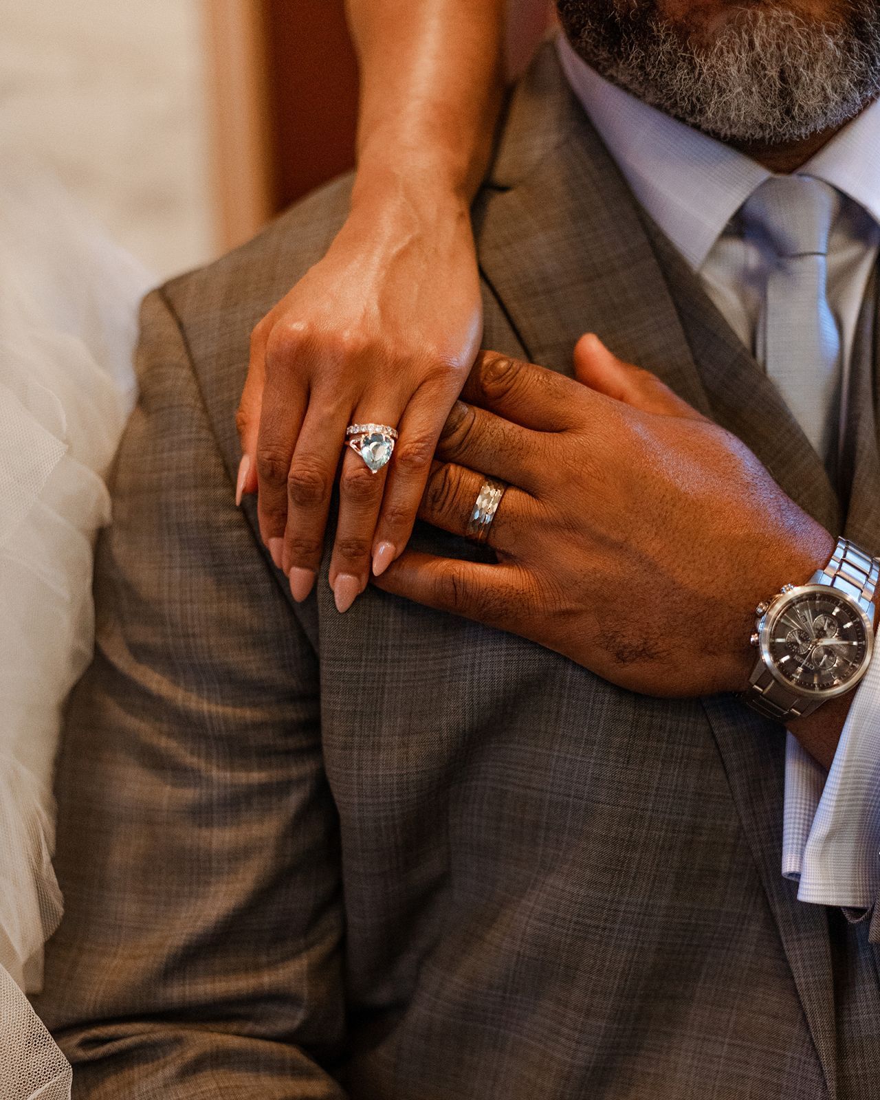 Close-up of bride's diamond engagement ring and groom's wedding band and watch on his gray plaid suit.