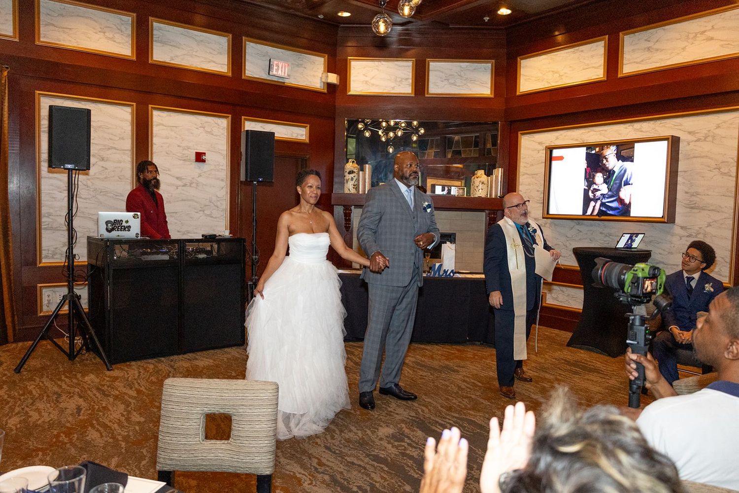 Bride and groom share first dance at elegant wedding reception with DJ and guests celebrating in background.
