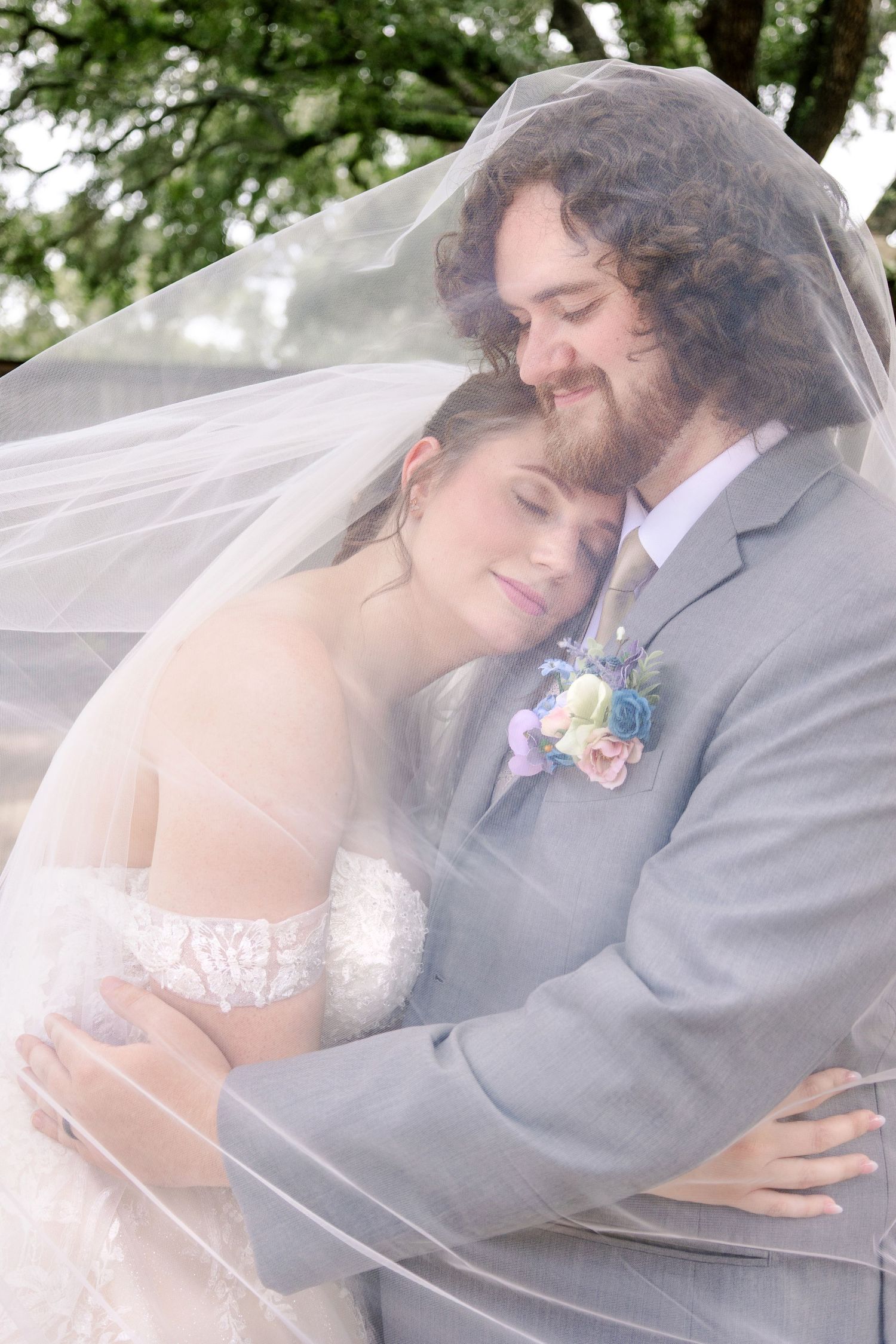 Romantic wedding couple embracing under a flowing veil in an outdoor setting.