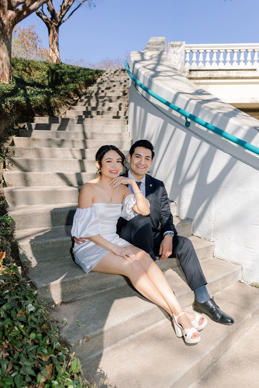 A couple in formal attire poses together on white stone steps in a romantic outdoor setting.