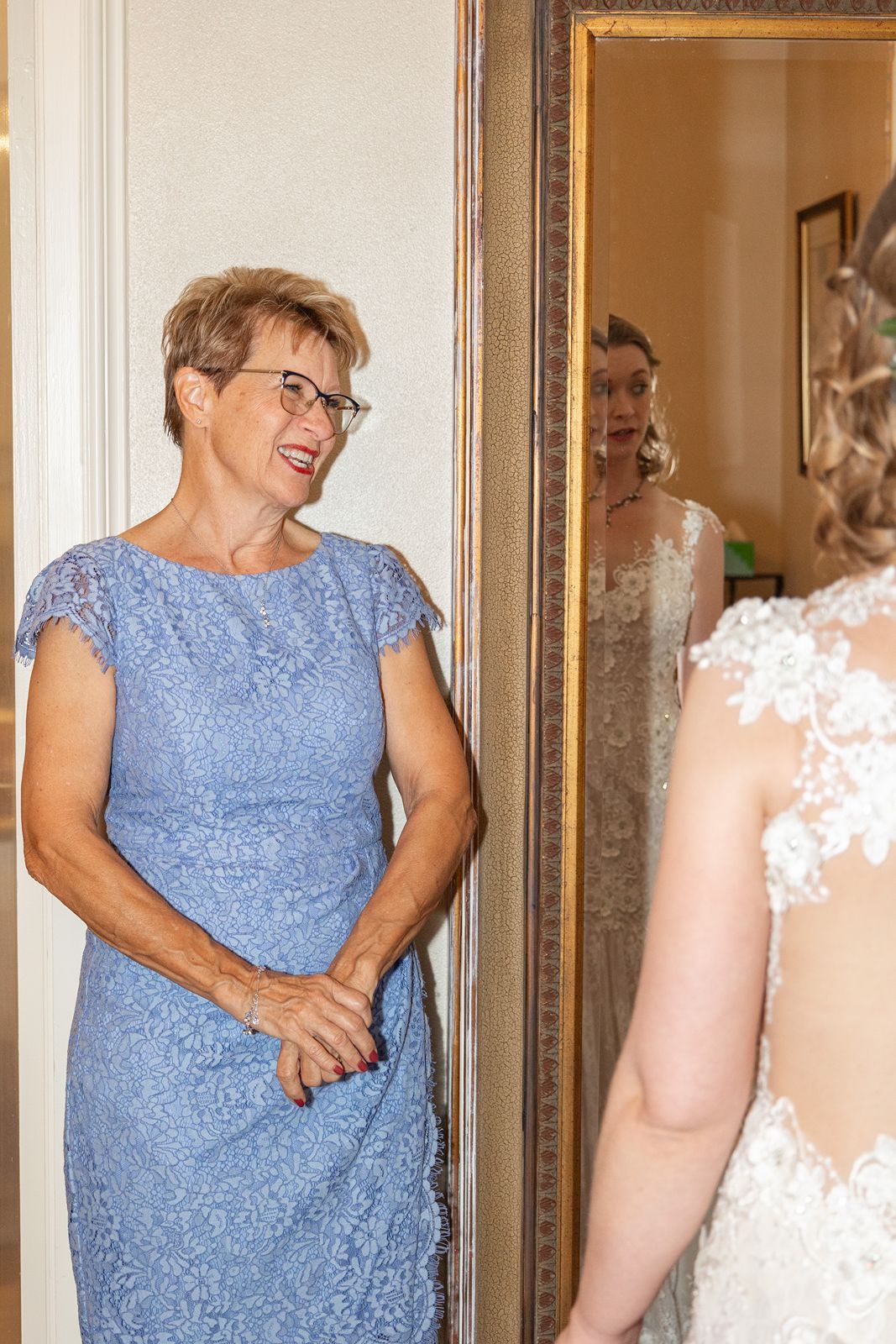A person in a light blue lace dress smiles while standing in front of ornate gold-framed mirrors.