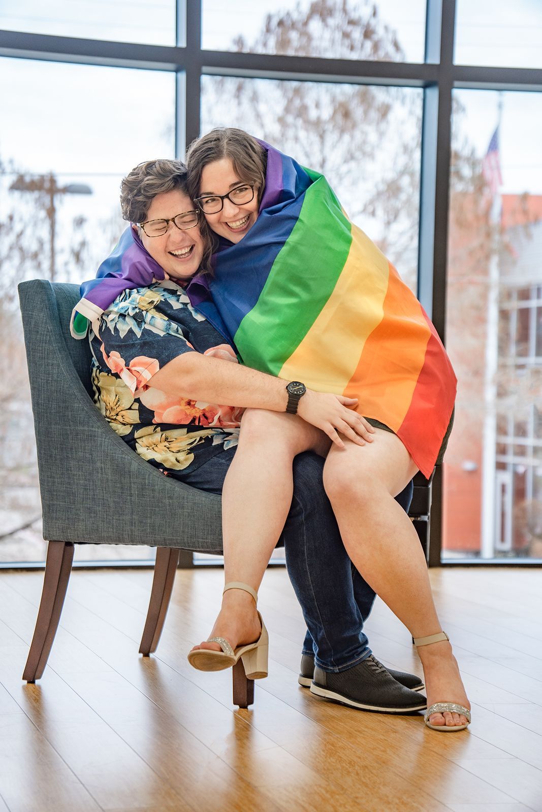 People sit in chairs holding a rainbow pride flag while laughing and smiling in a bright indoor space.