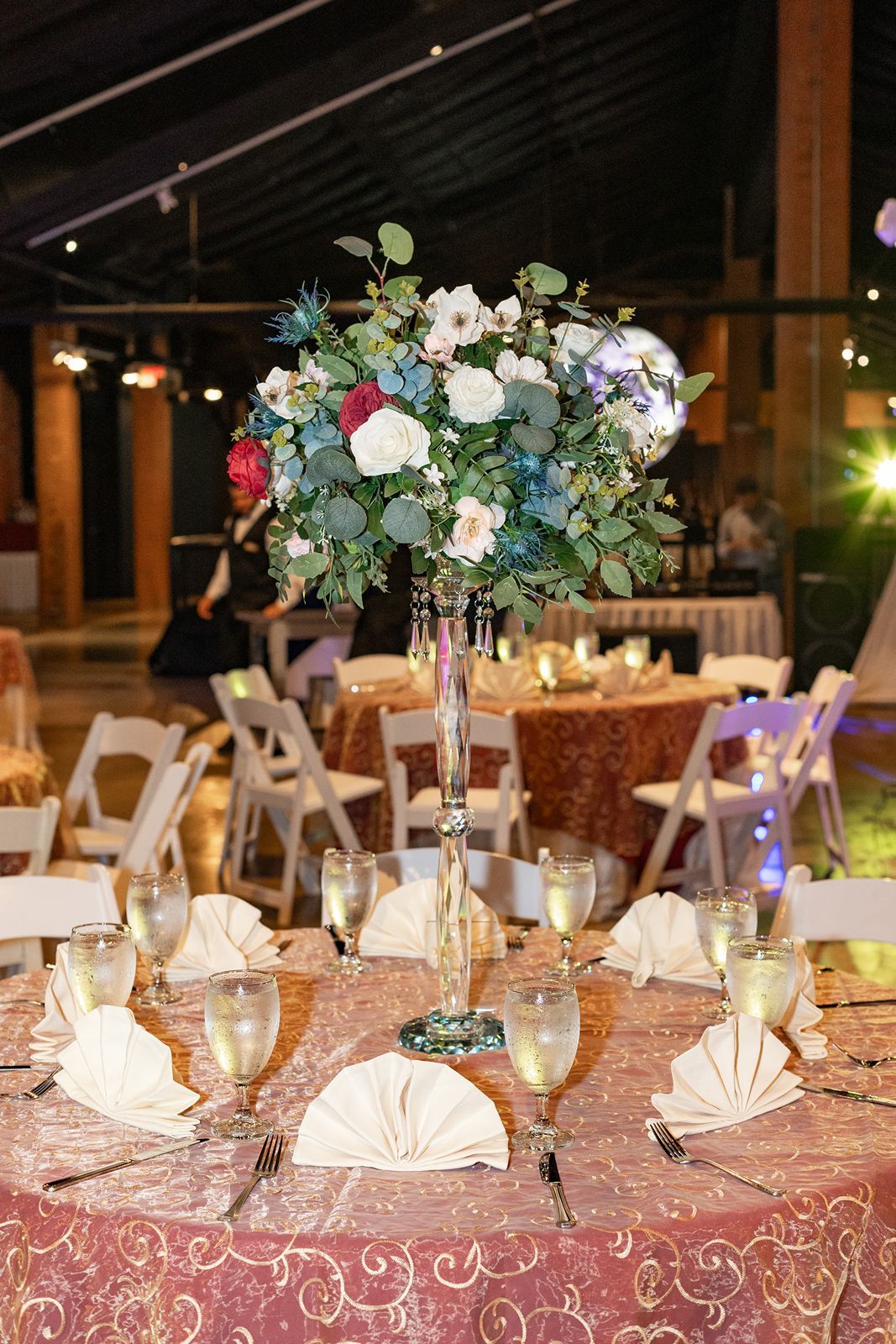 Elegant wedding reception table with tall floral centerpiece featuring white and burgundy roses on a pink embroidered tablecloth.
