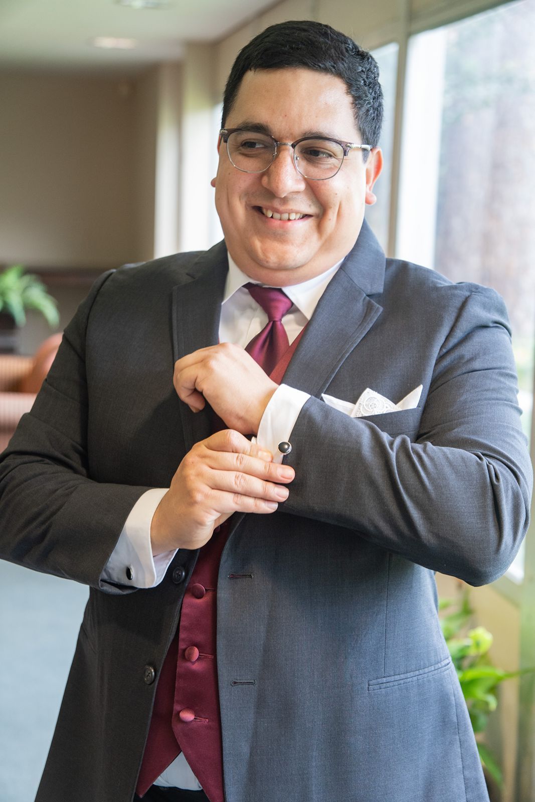 Groom in gray suit with burgundy vest adjusts cufflinks while smiling on wedding day.
