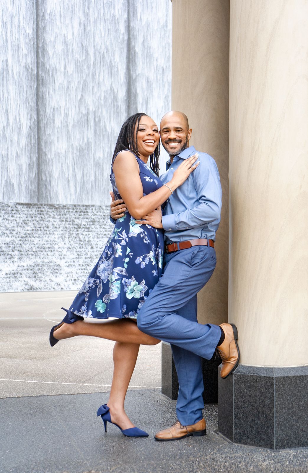 Couple in blue dress and gray suit embrace playfully against stone columns in romantic outdoor photo series.