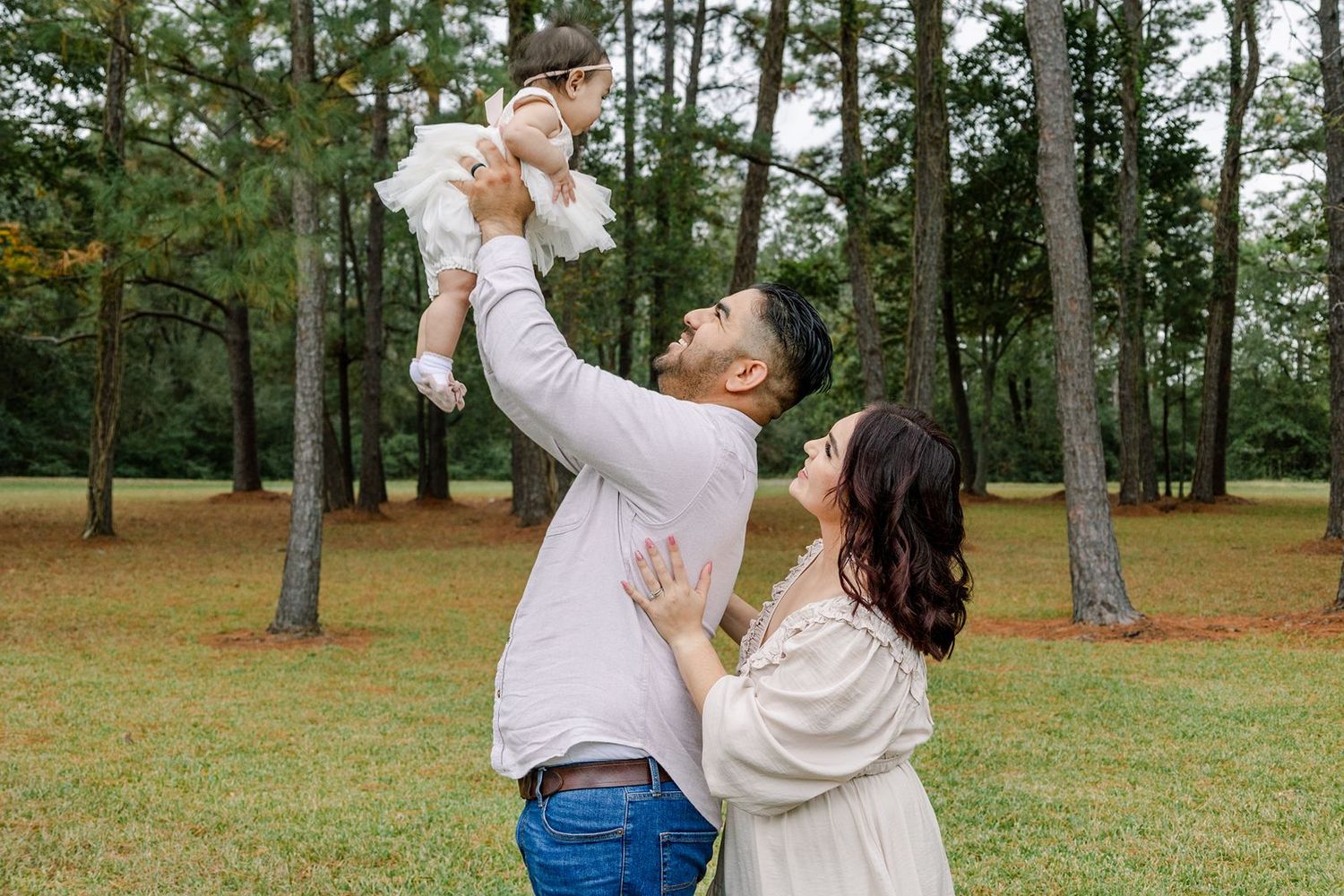 A couple dances together in a romantic embrace on a grassy lawn surrounded by tall pine trees.