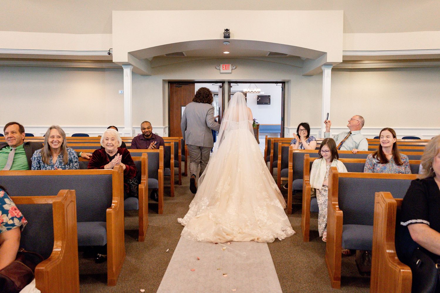 A bride in a white lace gown with flowing veil walks down the aisle of a church sanctuary with wooden pews.