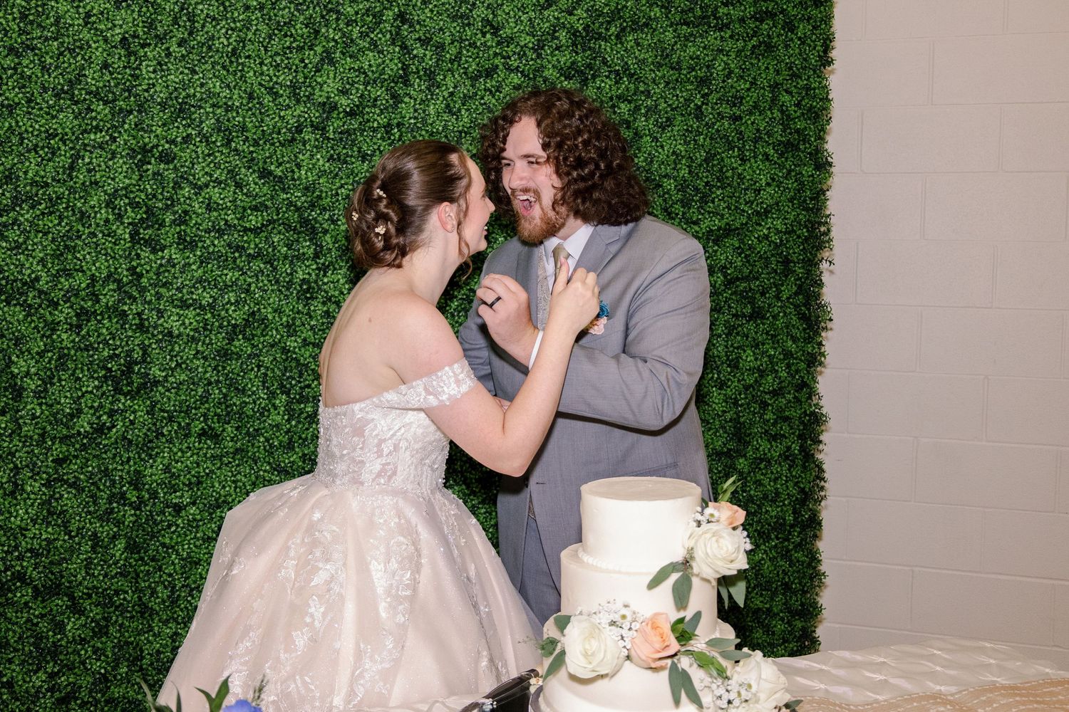 A couple cuts their elegant white wedding cake together against a green hedge backdrop.