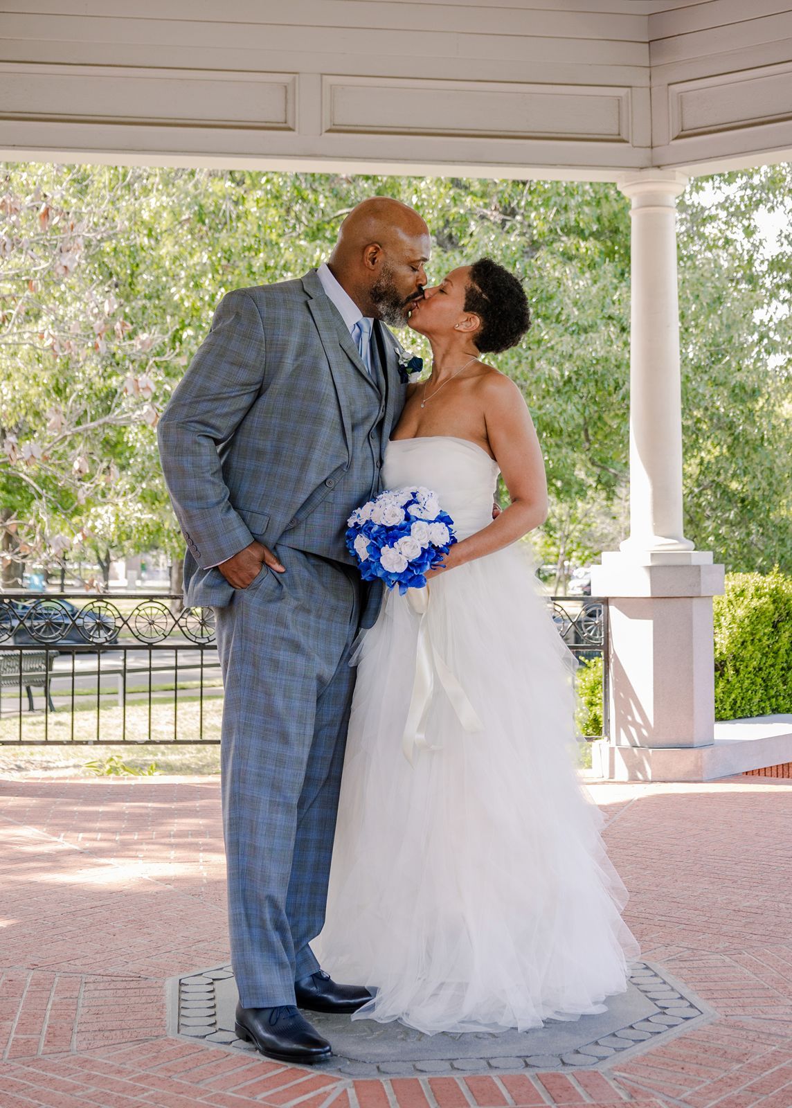 Wedding couple sharing a kiss under a gazebo, bride holding blue and white bouquet.
