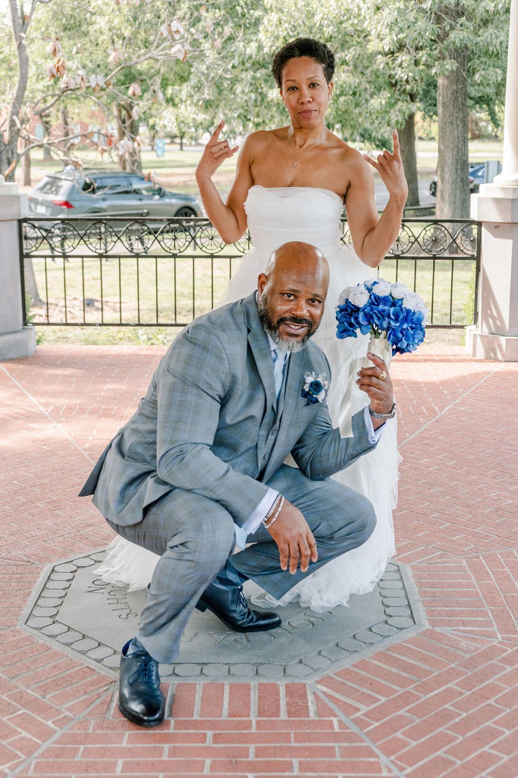 A bride and groom pose playfully outdoors, he crouches holding blue flowers while she stands behind making bunny ears.