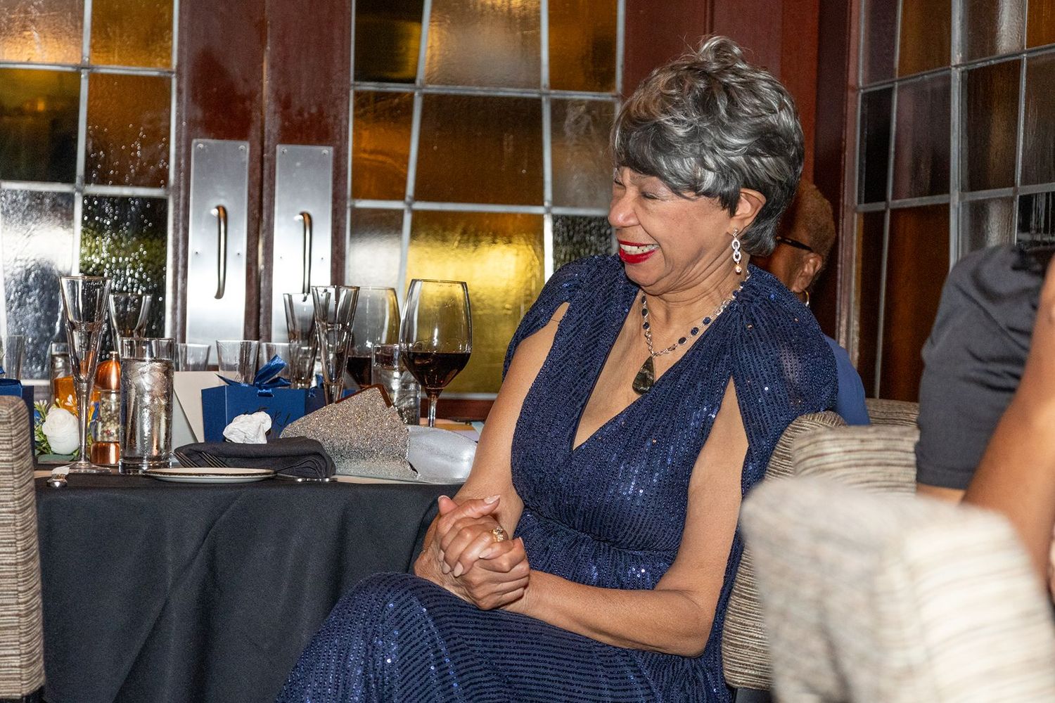 Elegant woman in navy blue sequin gown smiling at formal dinner event with wine glasses in background.
