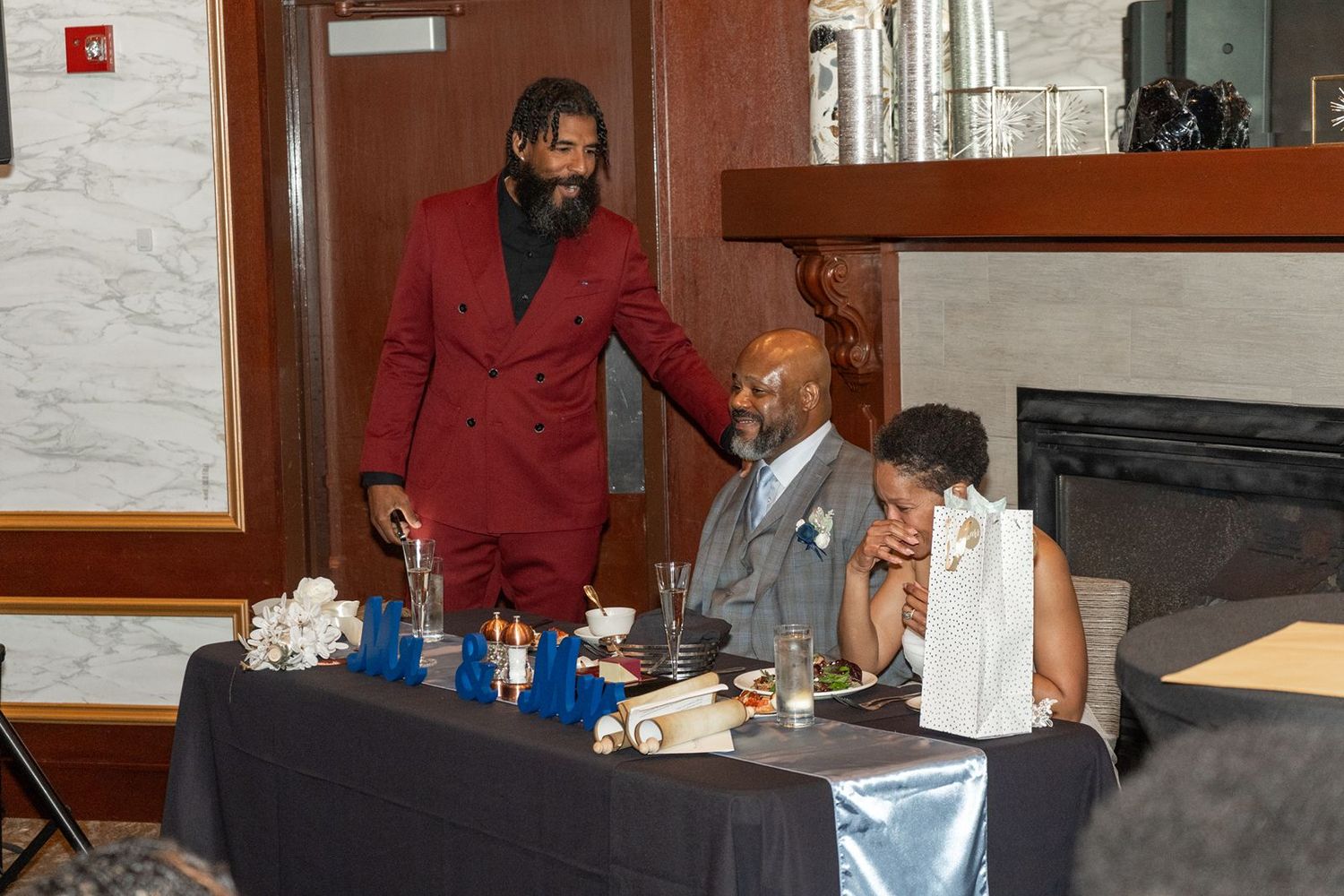 Man in red suit stands at sweetheart table speaking to seated couple at elegant wedding reception.