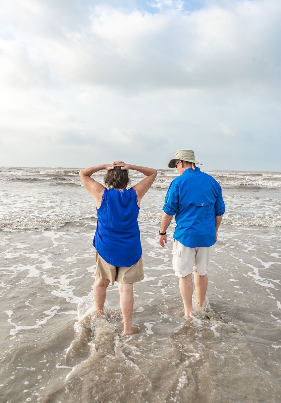 Two people in matching blue shirts walk along a beach shoreline as waves crash against the sandy coast.