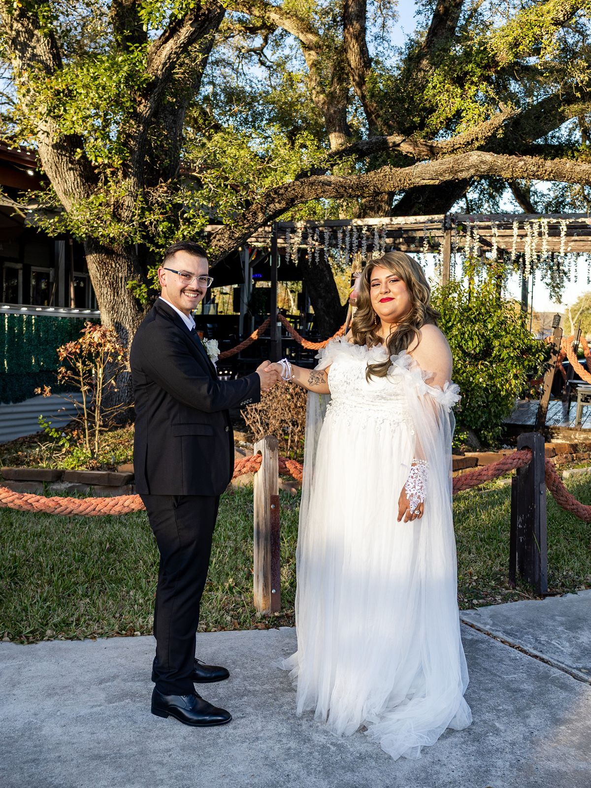 A couple in wedding attire shaking hands outdoors under oak trees, groom in black suit, bride in white gown.