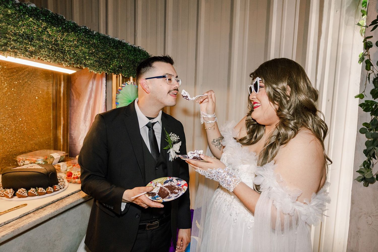Bride in white lace dress feeds groom in black suit wedding cake at elegant reception venue.