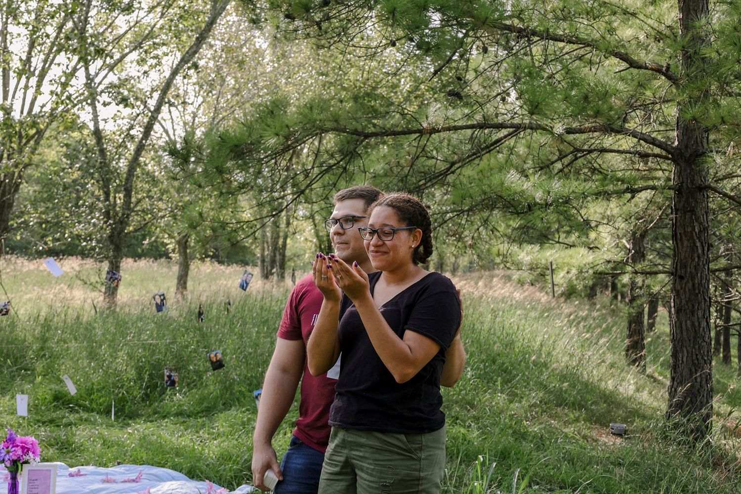 Two people clapping outdoors near trees and a table in a lush green meadow setting.