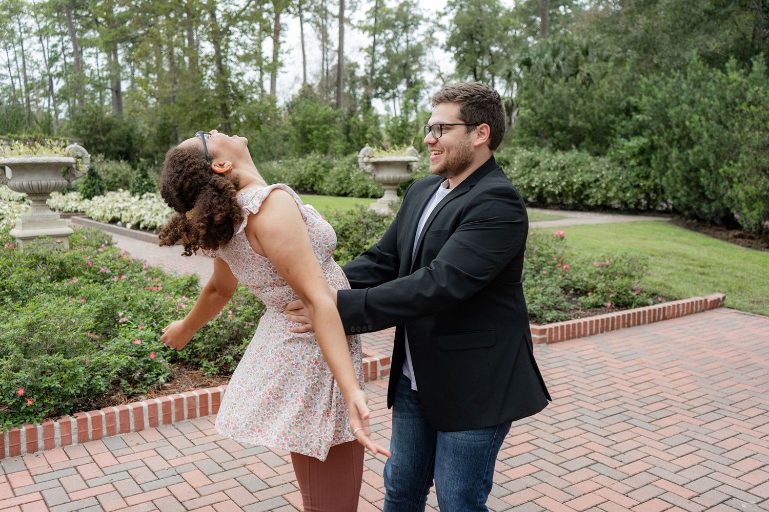 A couple dances together on a brick pathway in a lush garden, both wearing glasses and semi-formal attire.