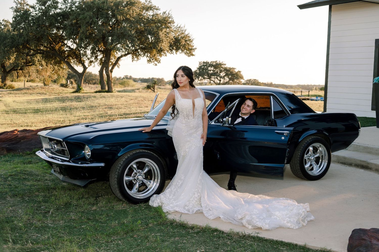 A classic black Ford Mustang sits in a field at sunset during a wedding photoshoot.