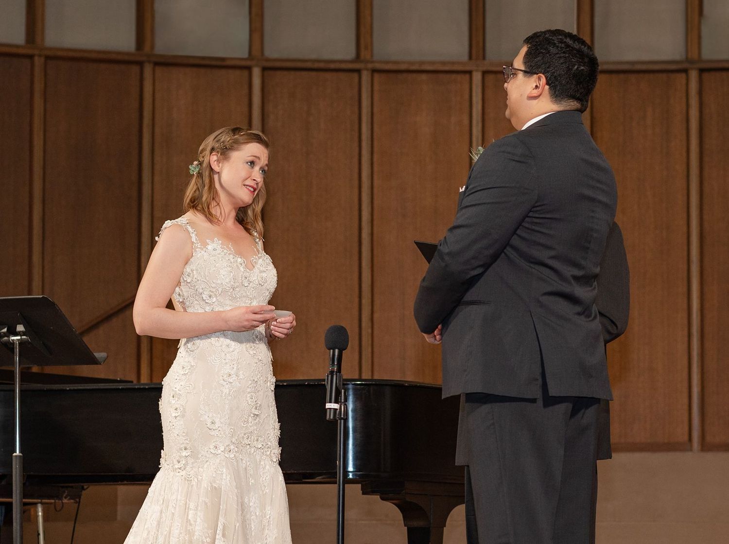 Bride in lace gown and groom in dark suit exchange vows on a concert hall stage.