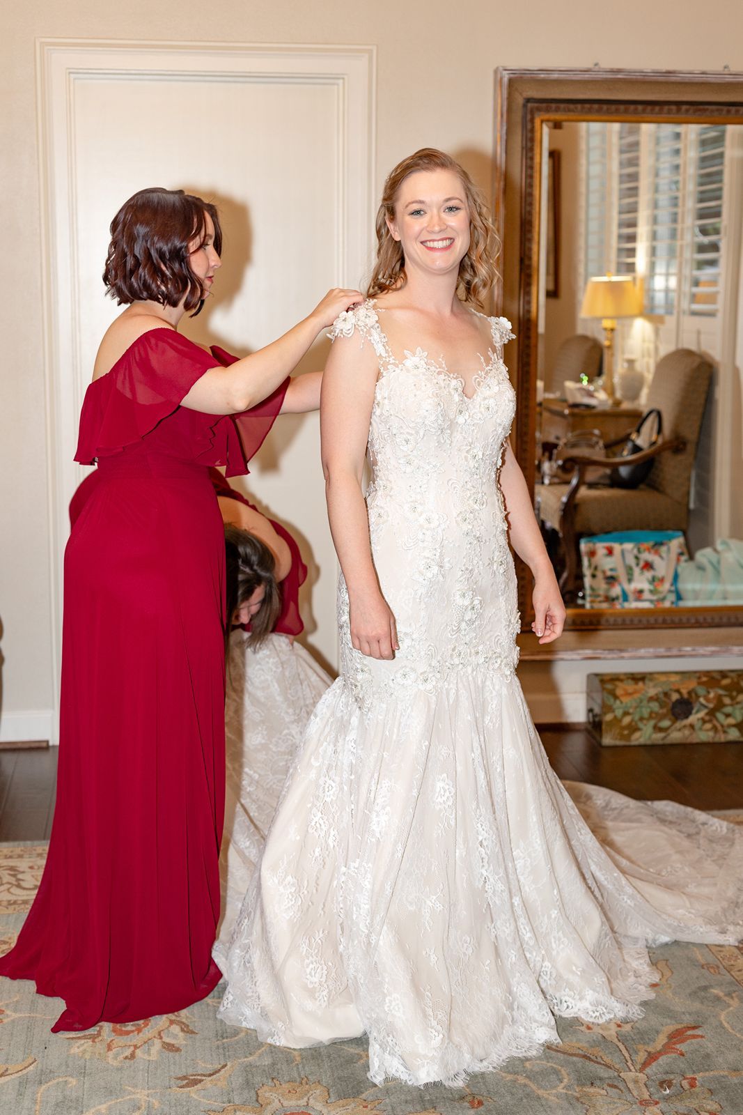 A bride in a white lace mermaid gown smiles as a woman in red adjusts her dress in an elegant room.