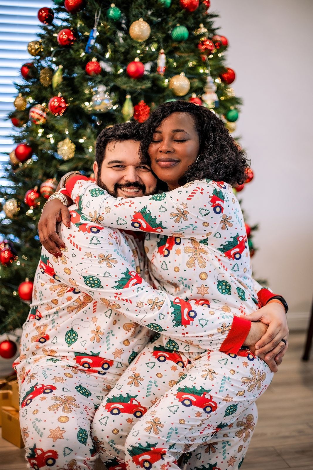 A couple wearing matching Christmas pajamas with red truck patterns hugs and laughs in front of a decorated tree.