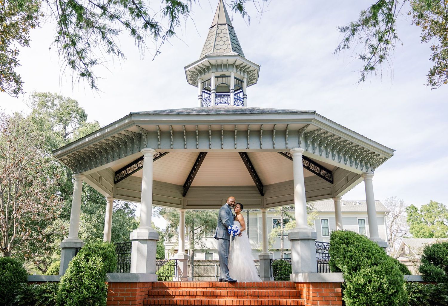 A white Victorian-style gazebo with ornate details and landscaping serves as a romantic wedding venue in a garden setting.