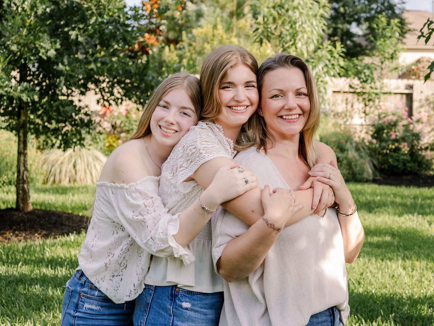 Three people embrace and laugh together in a sunny outdoor garden setting wearing light colored clothing.