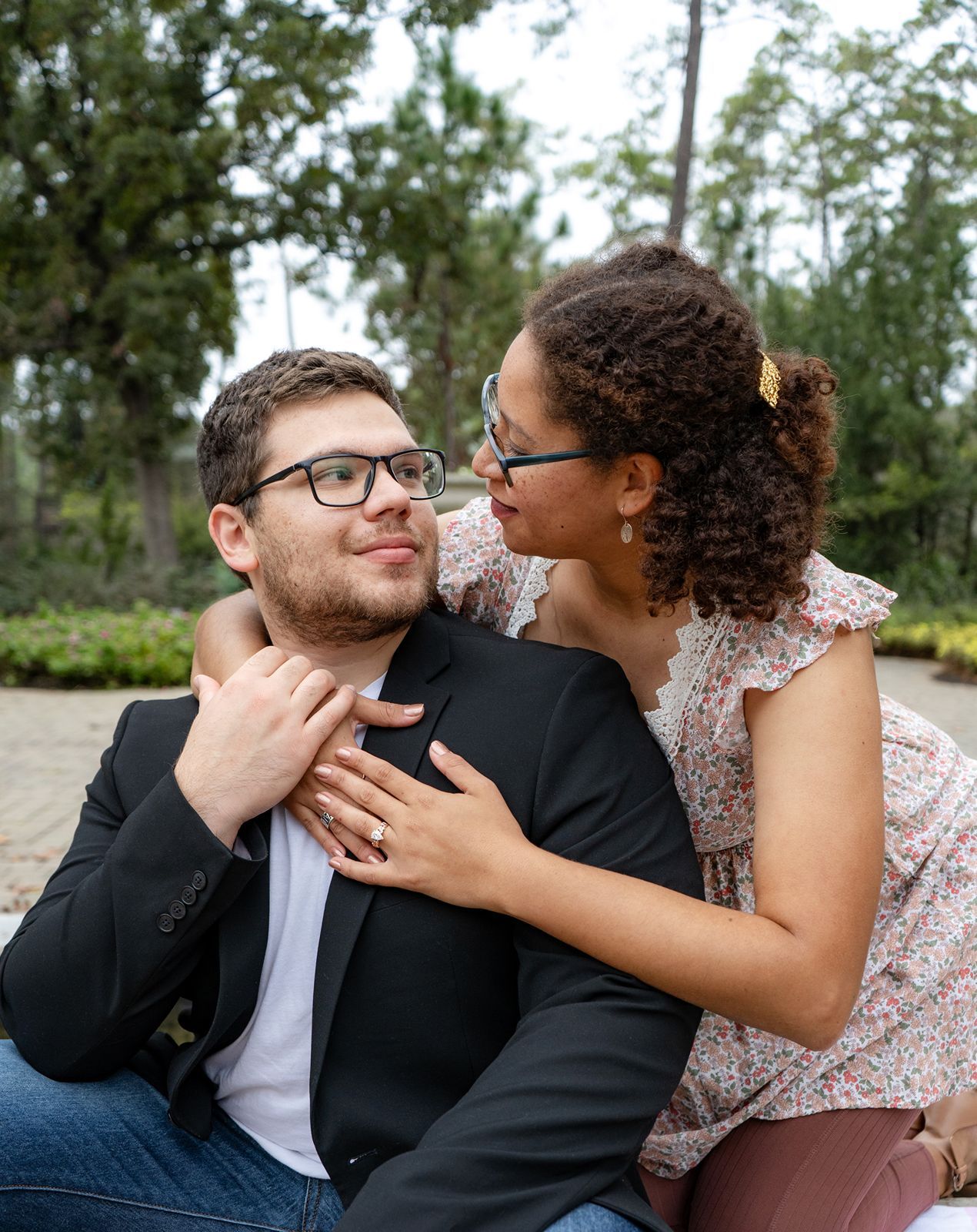 A couple shares a tender moment outdoors, woman in floral top embracing man in black blazer and glasses.