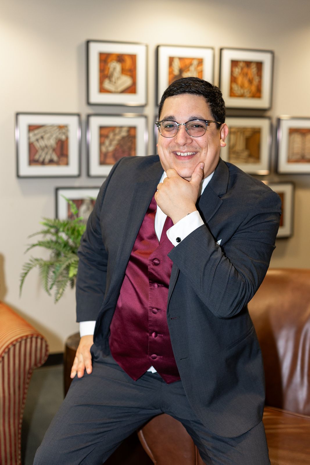 Smiling groom in gray suit and burgundy vest poses confidently in front of framed artwork.