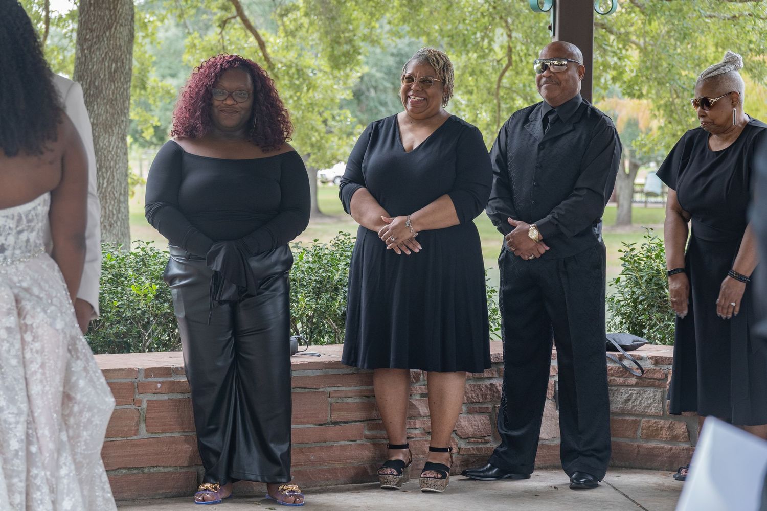 Wedding party members in black attire gather under a covered outdoor area during the celebration.