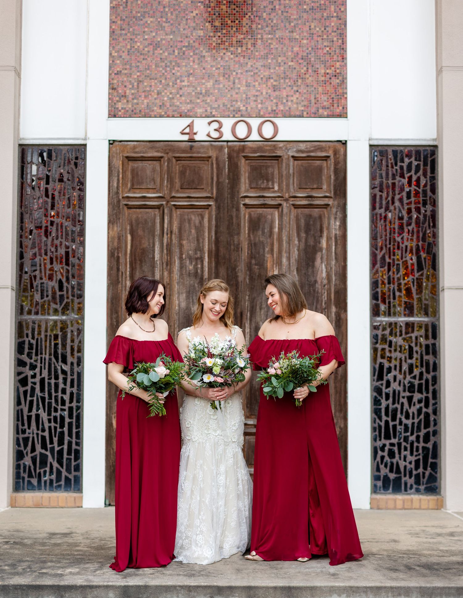 Bridal party in red dresses posing against church exterior with stained glass windows.