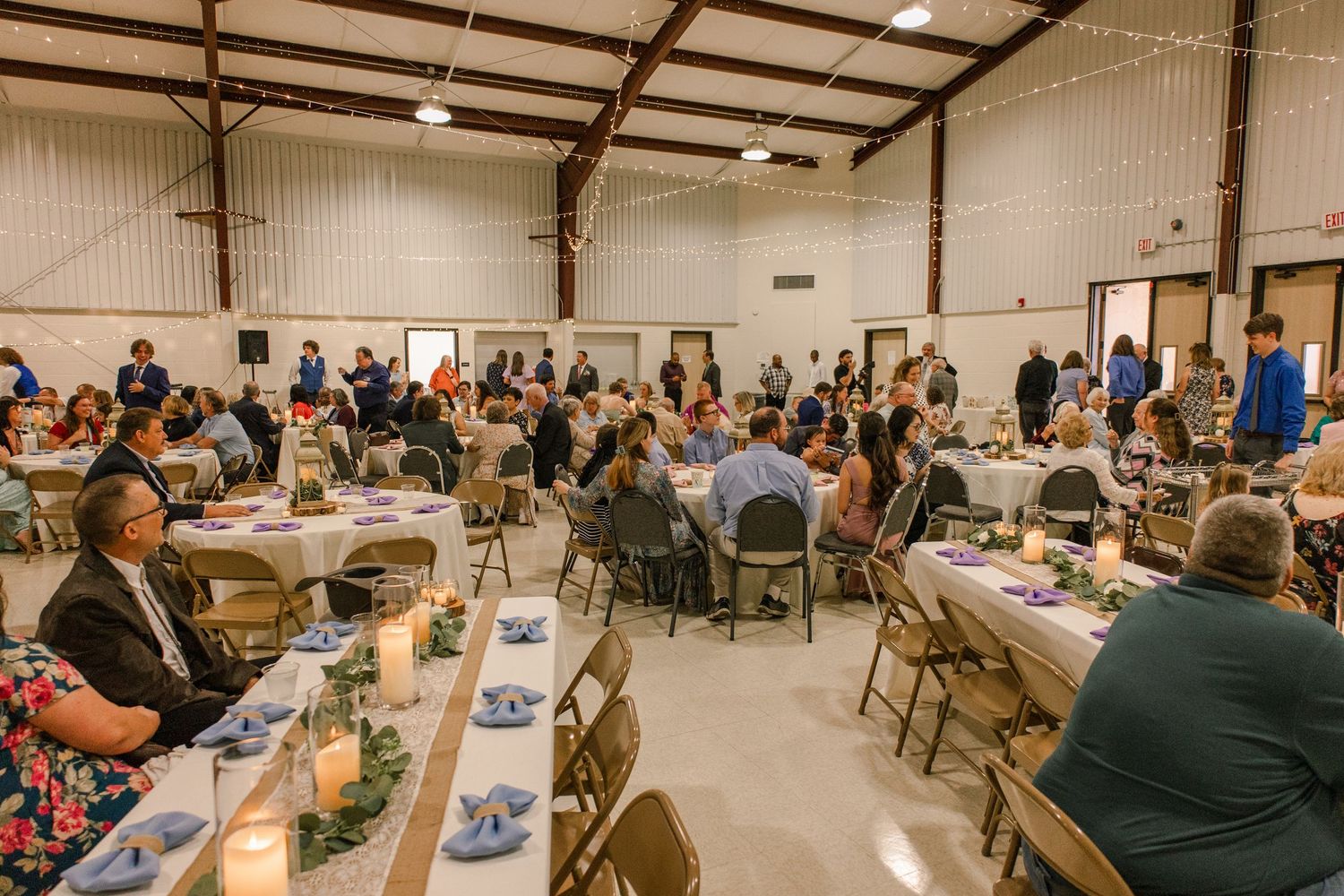 Large reception hall filled with guests seated at round tables decorated with string lights overhead.