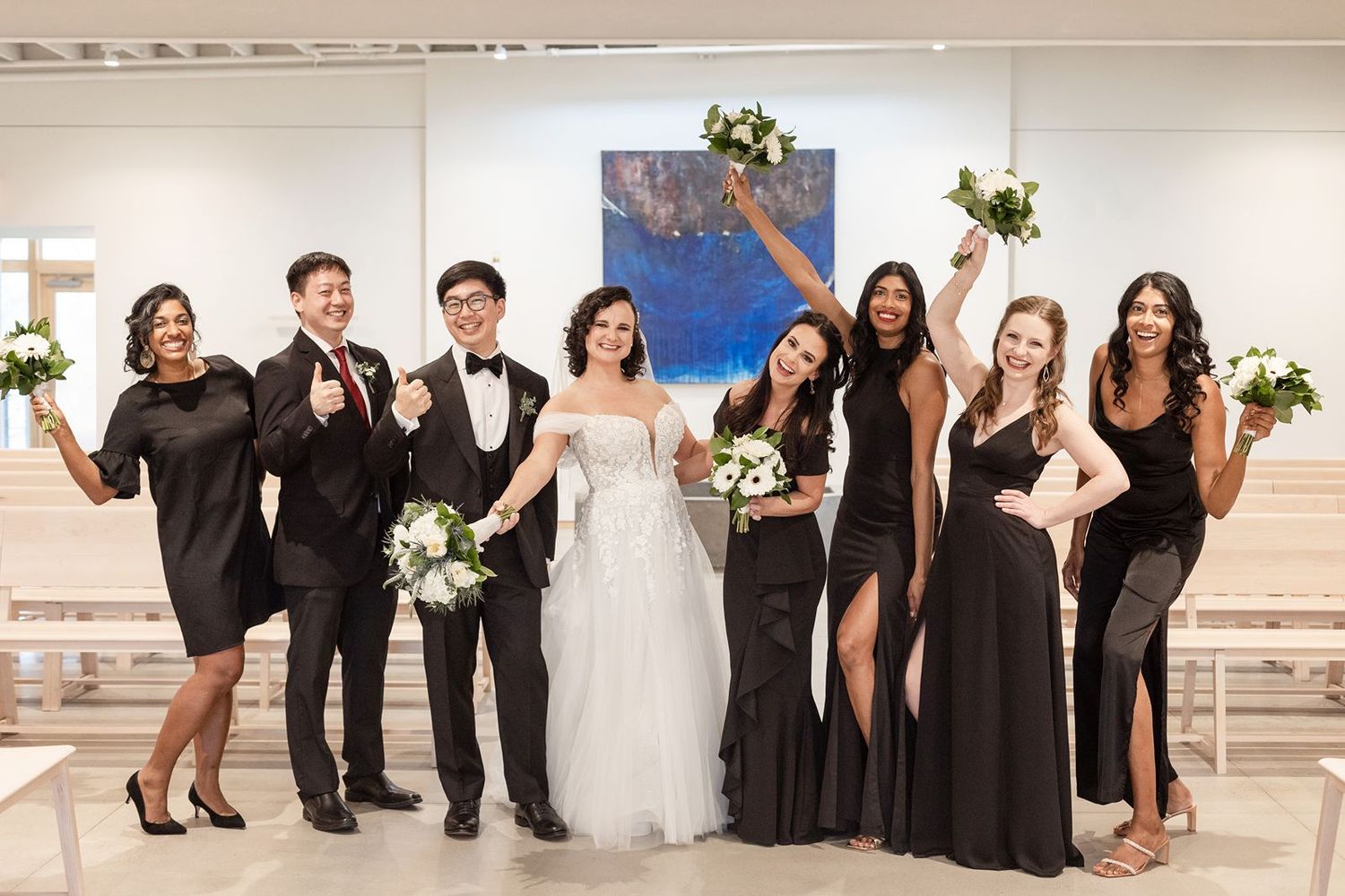 Wedding party members pose in a fun lineup wearing formal black and white attire at an indoor venue.