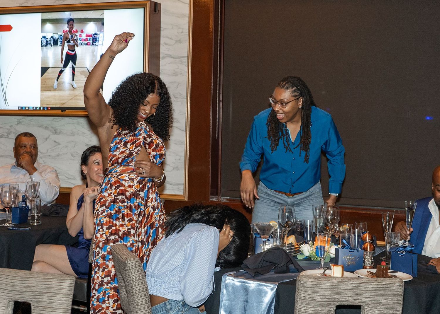 Women dancing and celebrating at an indoor event with tables, drinks, and a screen displaying a dance performance in the background.