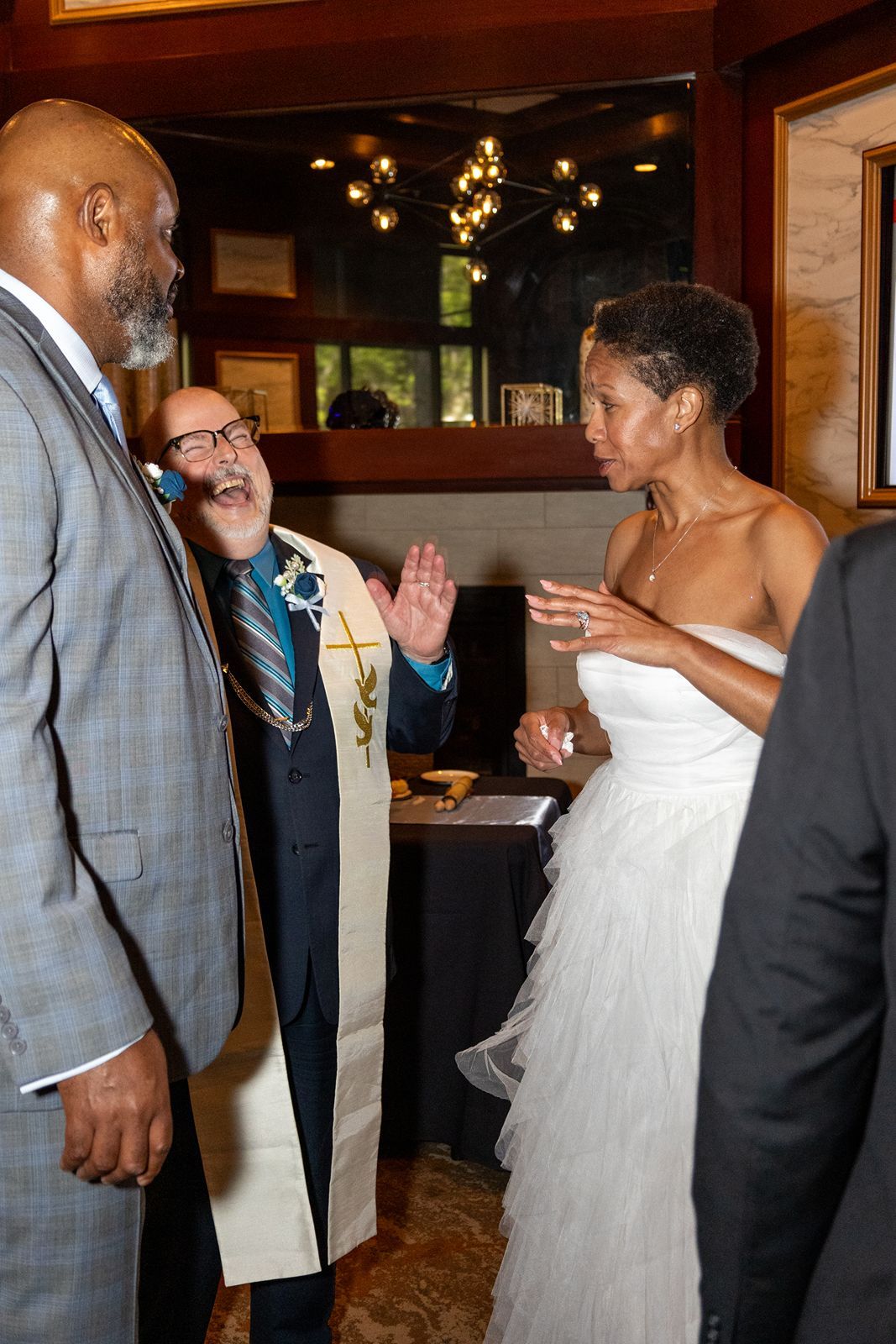 Officiant in ceremonial stole gestures while speaking with bride and groom during wedding ceremony.