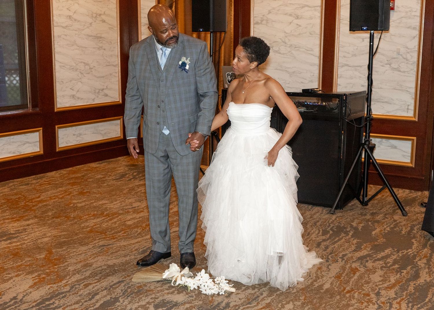Newlywed couple holding hands on dance floor, groom in gray plaid suit and bride in white tulle gown.