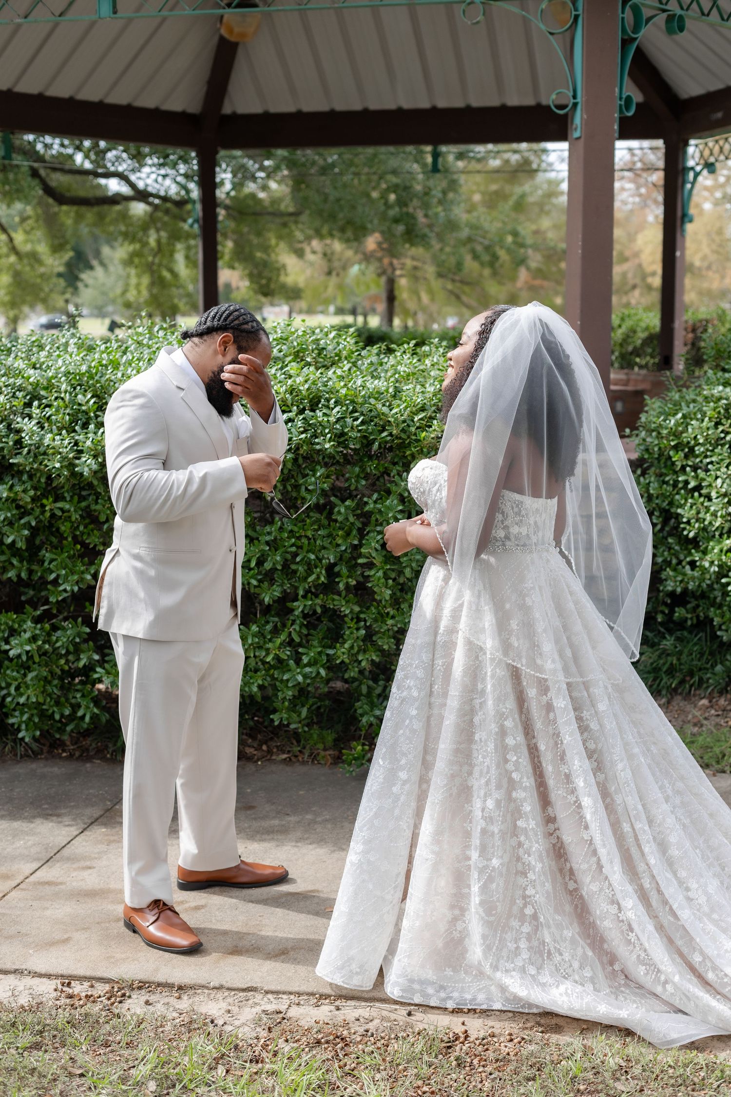 Groom wiping tears during emotional first look with bride in white lace gown and veil at outdoor gazebo.