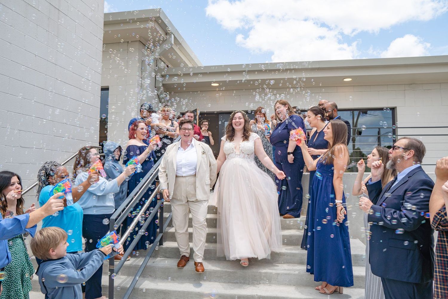 Newlywed couple exits wedding venue on steps as guests celebrate with bubbles in blue dresses and suits.