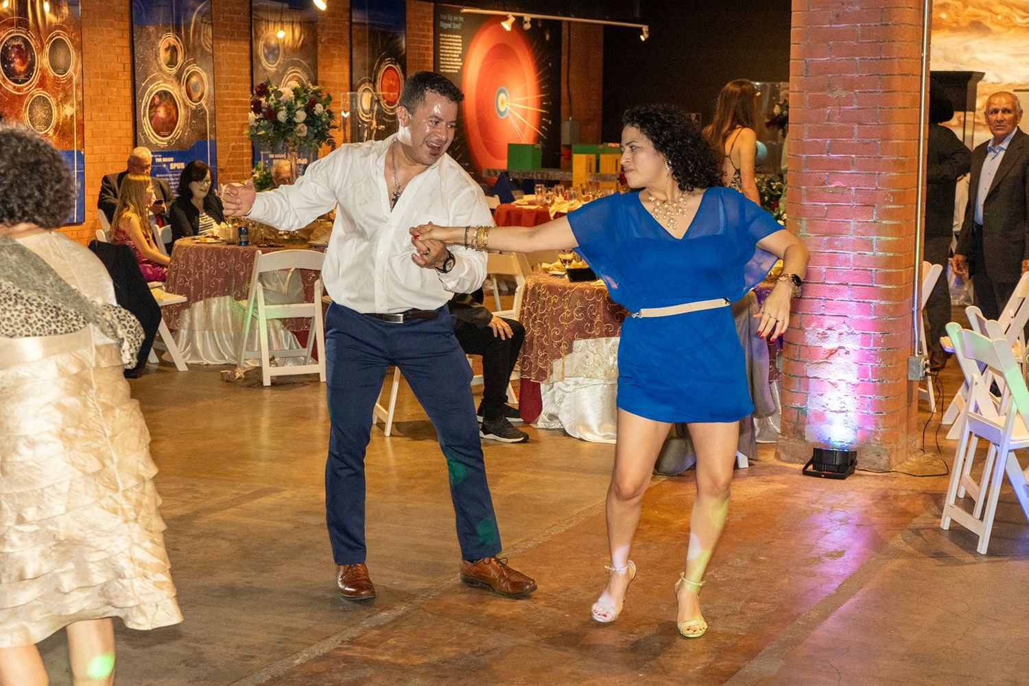 A couple dances energetically at a wedding reception, woman in a blue dress and man in a white shirt.