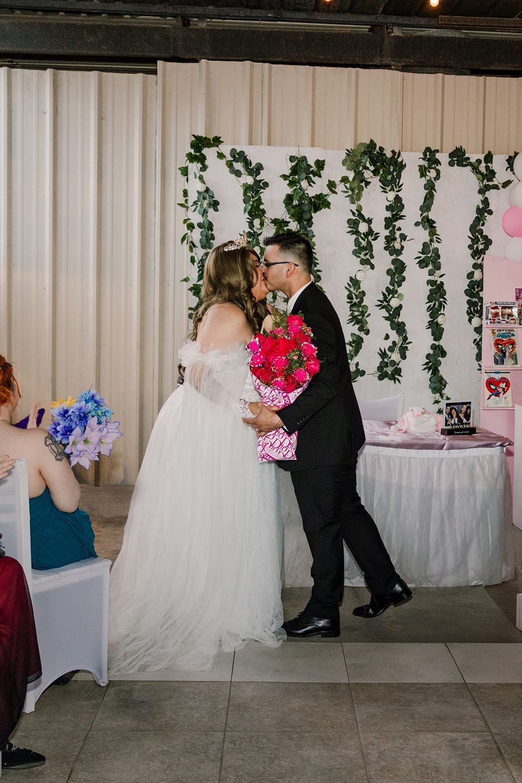 Bride and groom share a kiss at wedding reception with greenery backdrop, bride holds pink bouquet, groom in black suit.