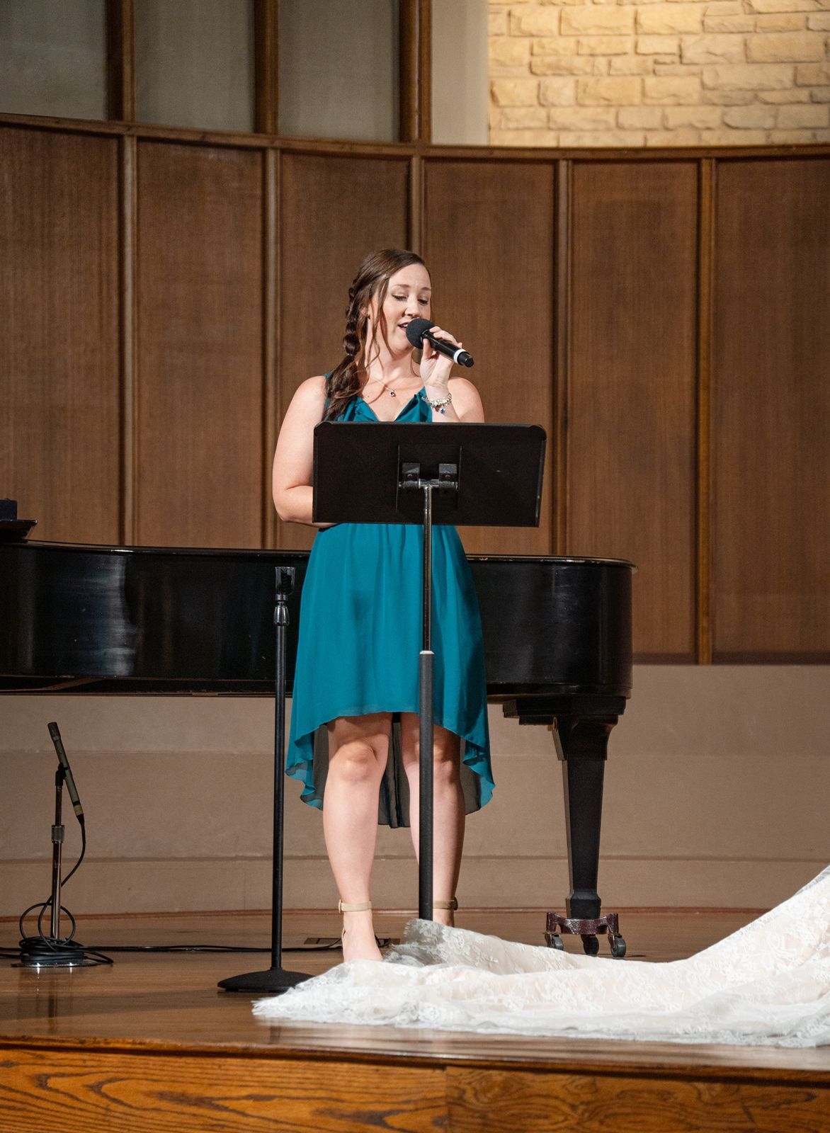 A performer in a teal dress plays piano in a recital hall with wood paneling and brick walls.
