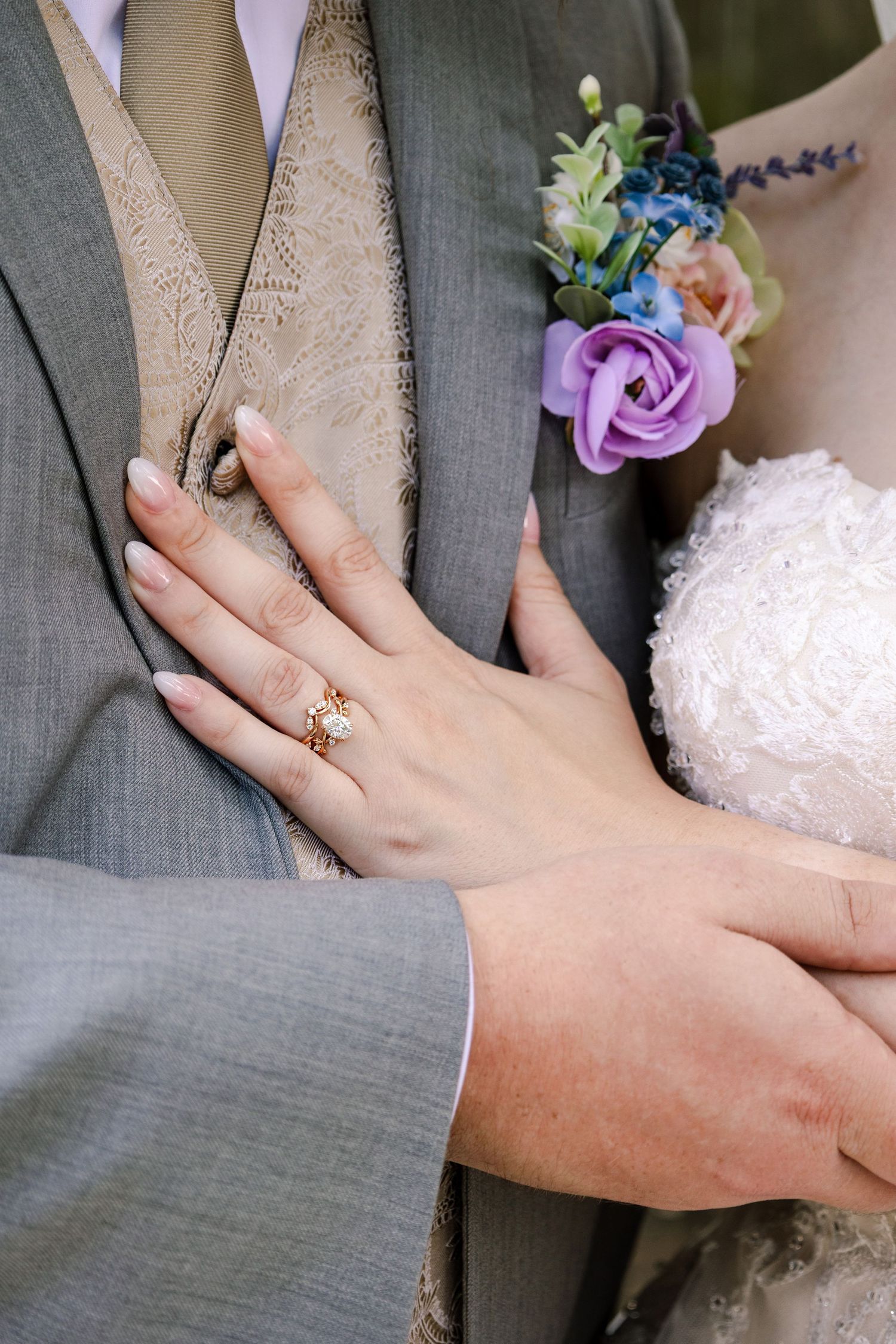 Close-up of hands showing an engagement ring with a colorful floral boutonniere on a grey suit.