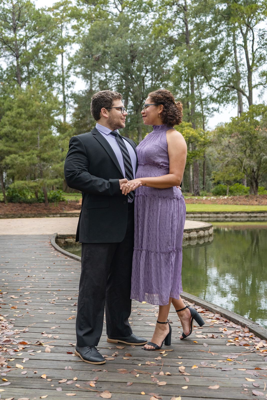 Couple in formal attire holding hands on a wooden dock by a pond surrounded by tall trees.