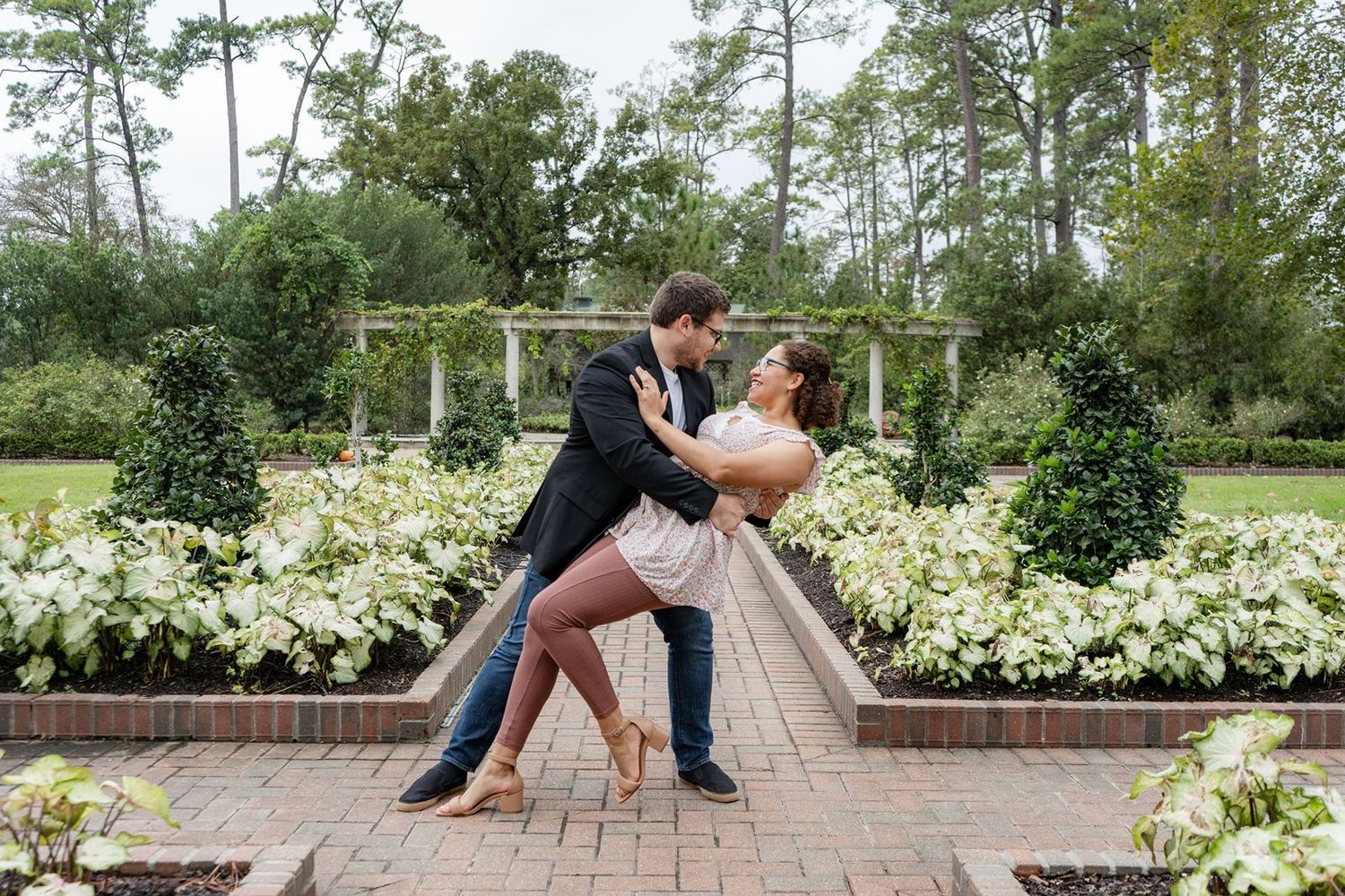 Couple dancing romantically in a lush garden with white flowers and brick pathway during an engagement photo session.