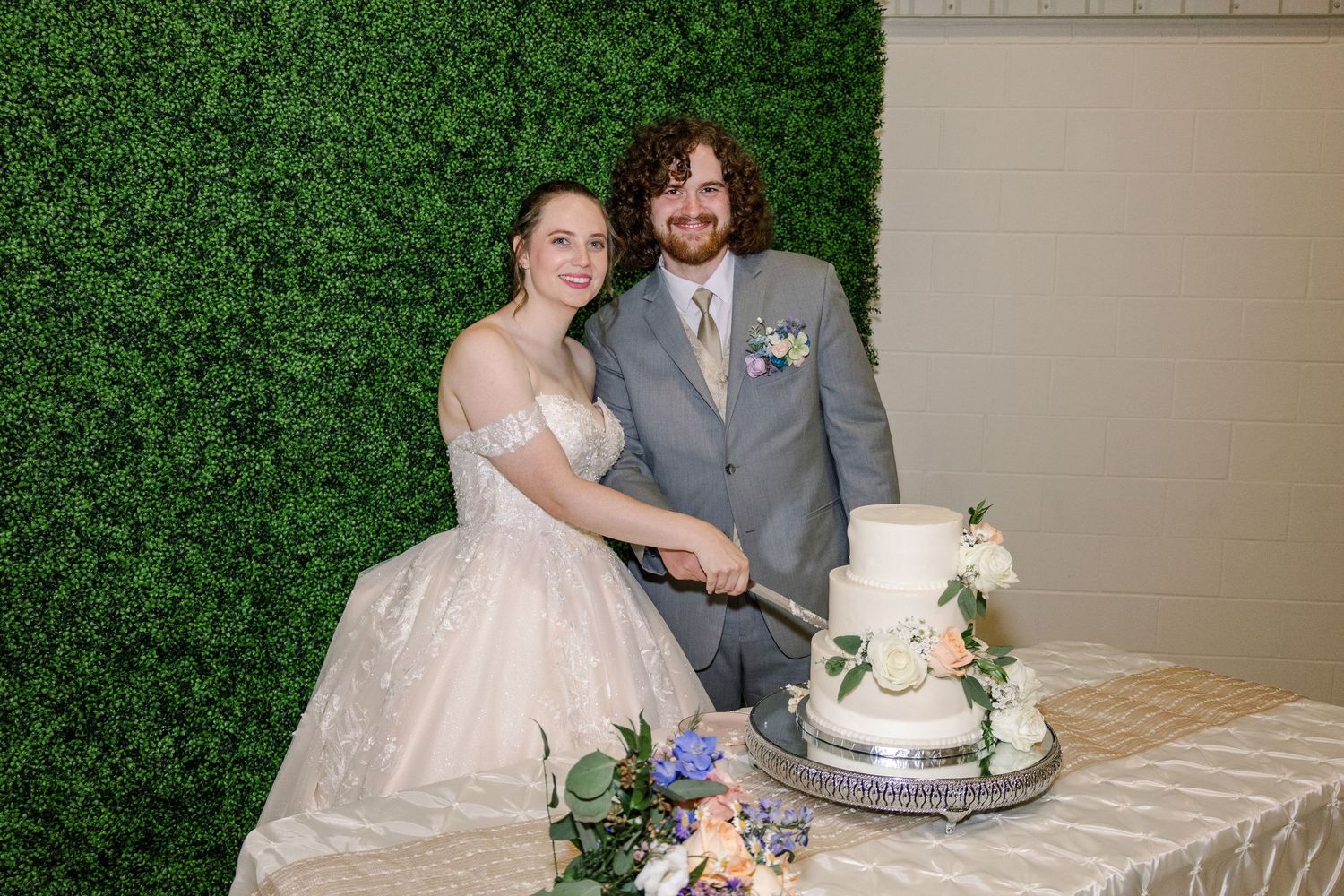 Newlyweds pose beside their three-tiered white wedding cake with floral decorations.