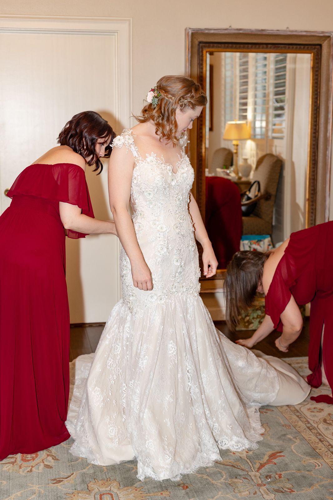 Bridesmaids in red dresses help bride into elegant lace mermaid wedding gown in a classic interior room.