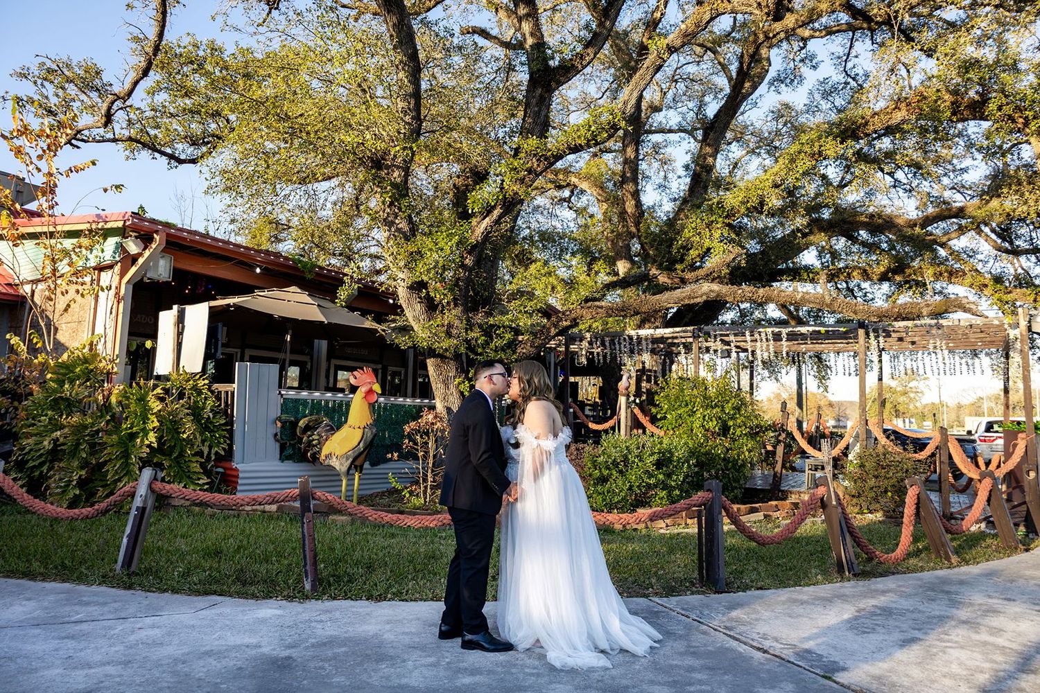 Bride and groom share a kiss outdoors under a large oak tree at a rustic wedding venue.
