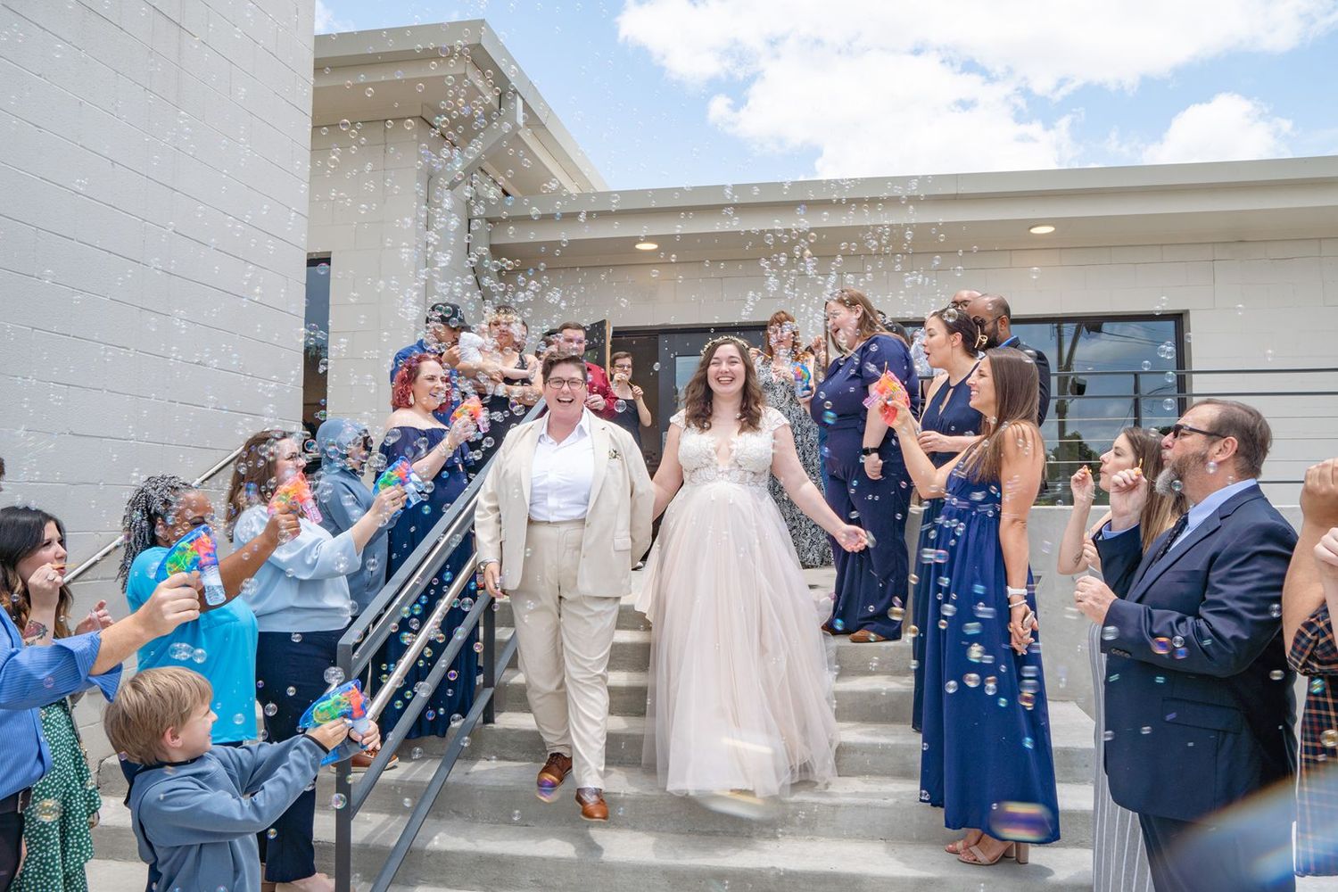 Wedding guests blow bubbles to celebrate the newlyweds outside a white stone building on a sunny day.