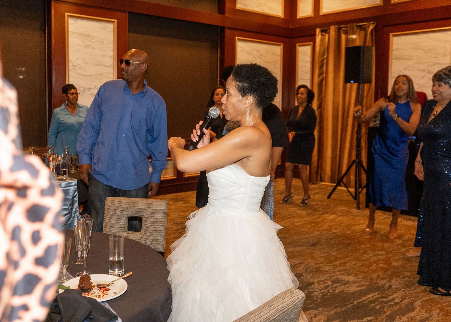 Bride gives speech with microphone at elegant wedding reception surrounded by guests.