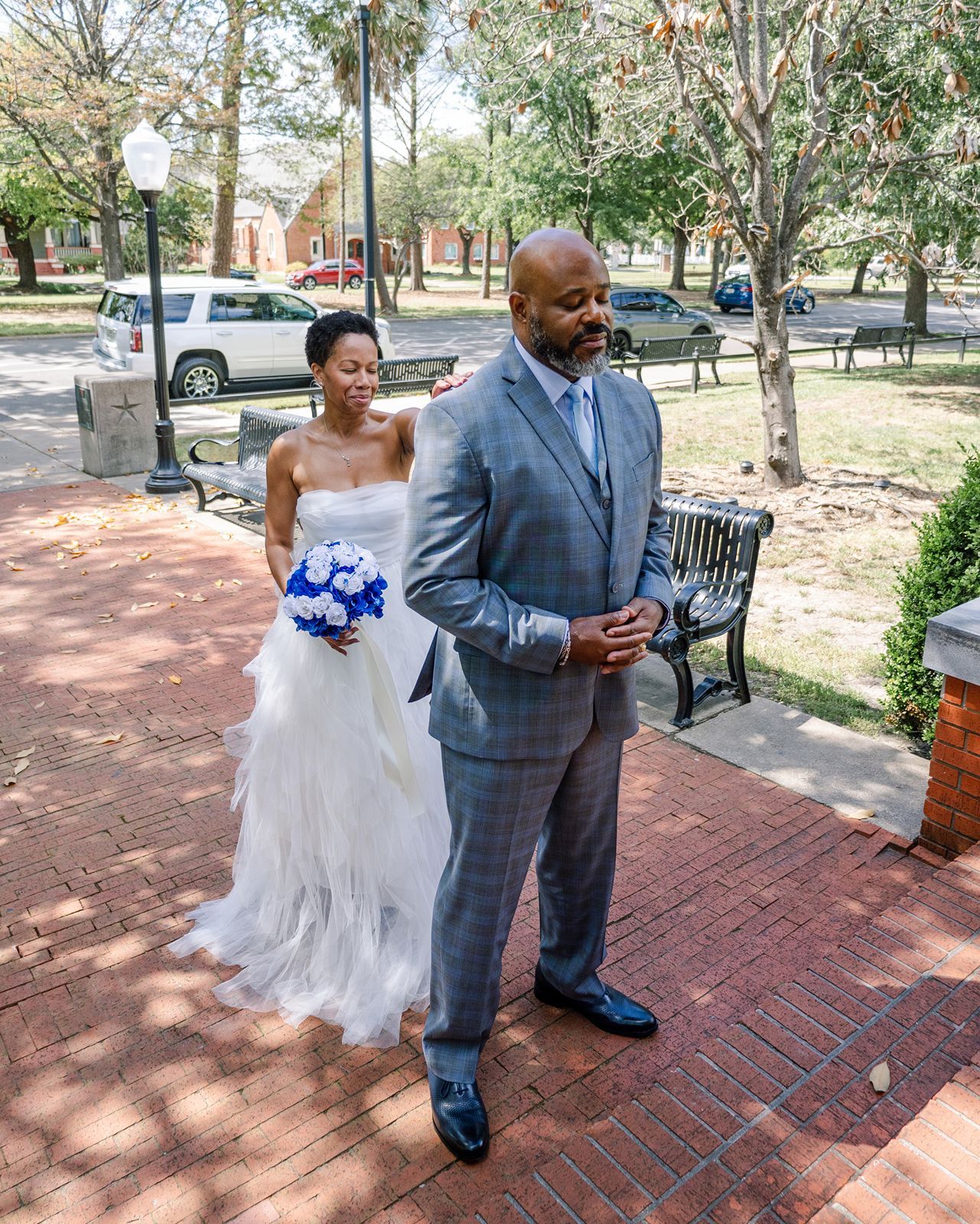 Bride in strapless white gown and groom in gray suit share emotional first look on brick pathway.